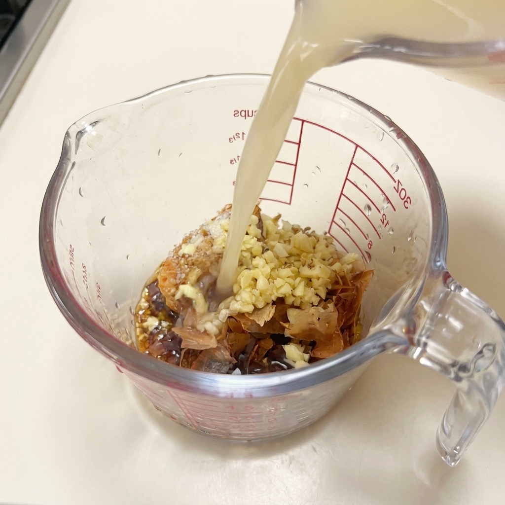 Hot, opaque pork bone broth being poured from a pitcher into a measuring cup filled with miso paste, garlic, bonito flakes, and liquid seasonings.