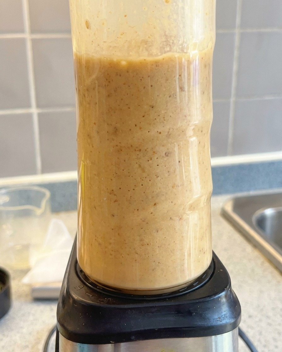 A tall food processor blending a creamy, light brown tonkotsu soup base until smooth.