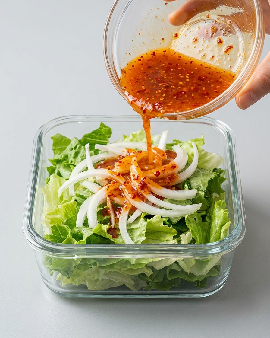 Pouring a vibrant red, spicy kimchi broth from a small glass bowl over fresh lettuce and sliced white onions in a rectangular glass container.