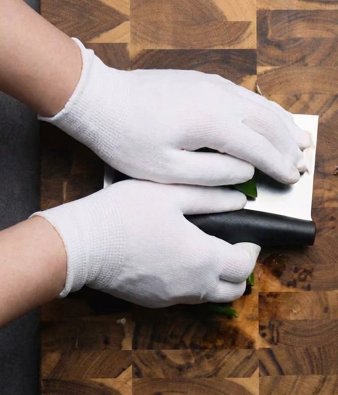 Gloved hands using a cleaver to chop green chilies on a wooden cutting board.