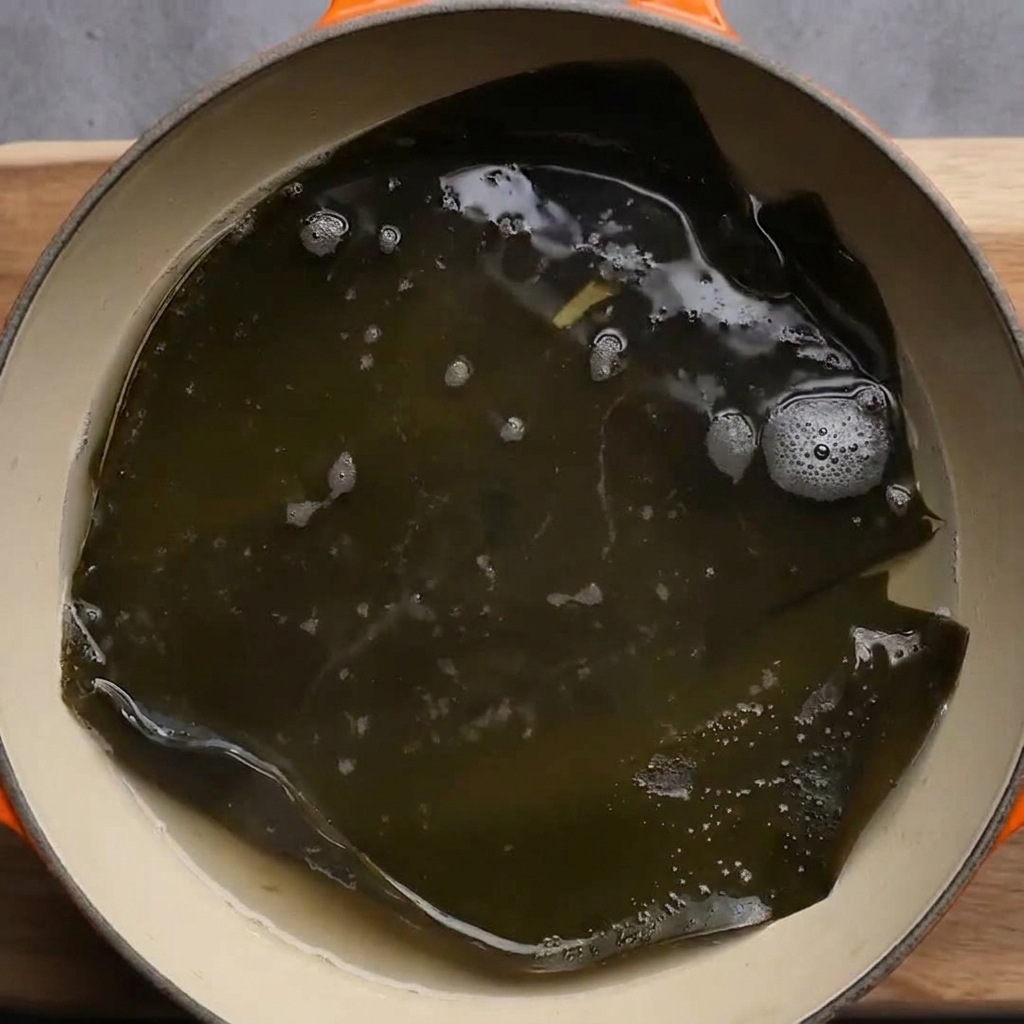 A large, dark green piece of dried kelp simmering in a pot of bubbling hot water.