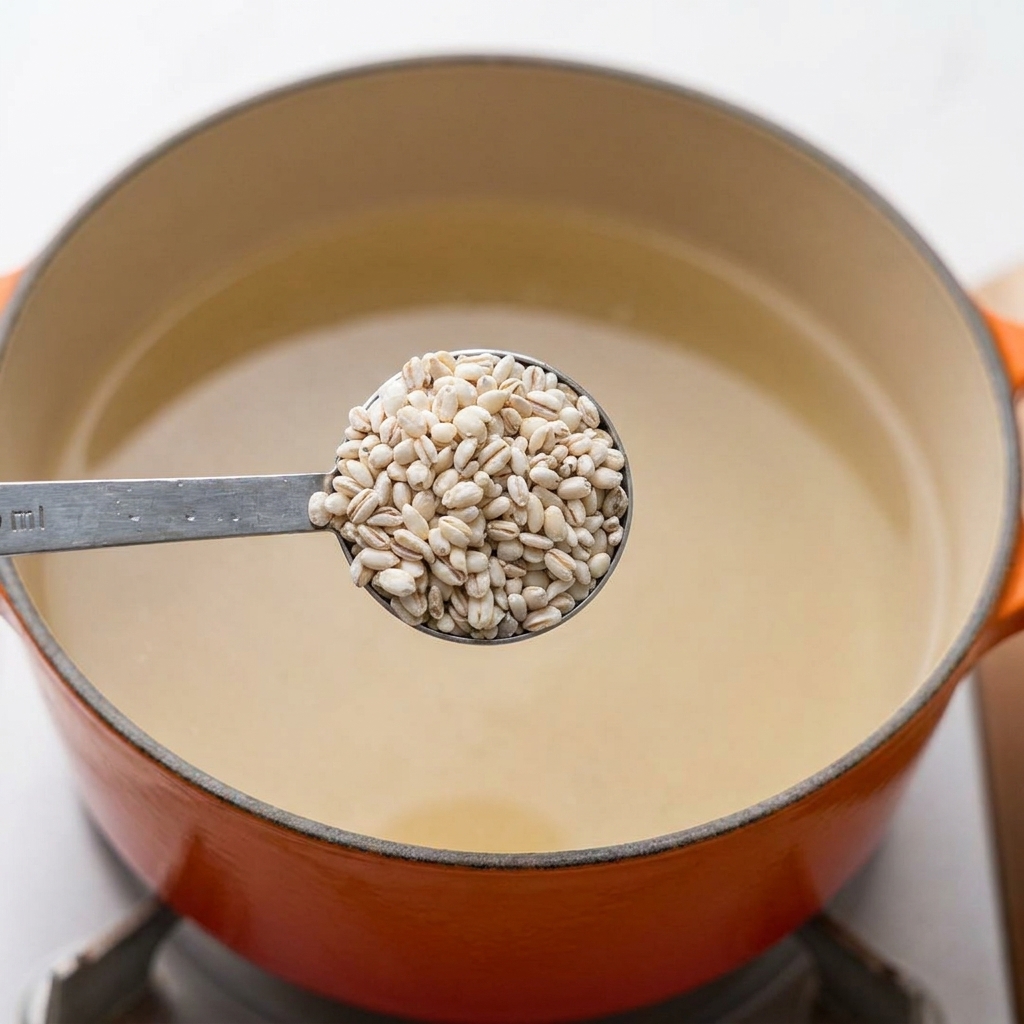 A metal measuring spoon filled with whole raw barley grains held directly over a pot of boiling water.