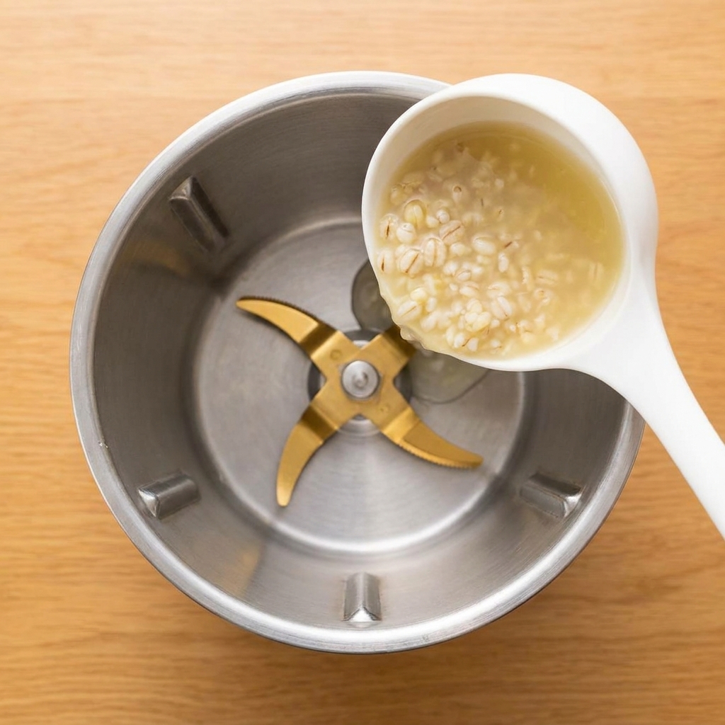 A white ladle pouring a thick, pale barley porridge into a stainless steel blender jar.