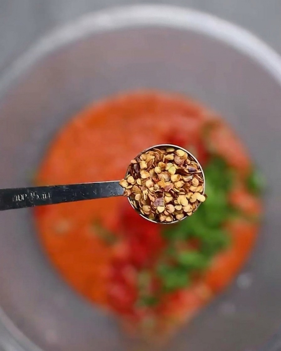 A metal measuring spoon holding dry chili seeds, held closely over a bowl containing blended red chili paste and fresh chopped vegetables.