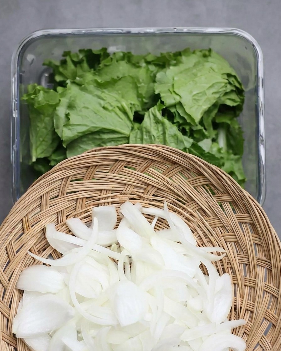 A woven basket of thinly sliced white onions held above a glass container filled with fresh green lettuce leaves.