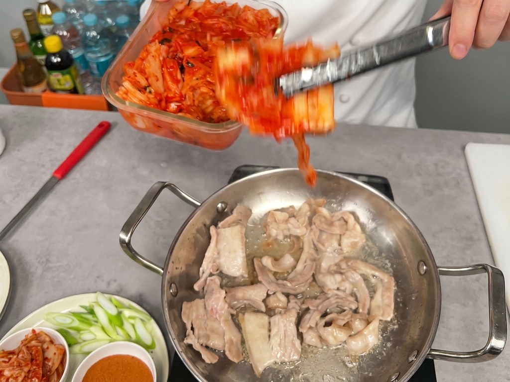 A large portion of bright red kimchi being transferred from a glass container into a pan with partially cooked pork belly slices.