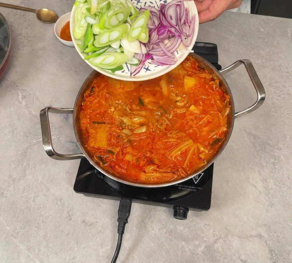 Sliced green onions and red onions being slid off a patterned plate into a simmering pot of red kimchi stew.