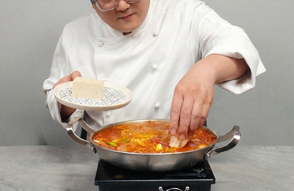 A person placing slices of firm white tofu into a pot of hot, bubbling kimchi stew.