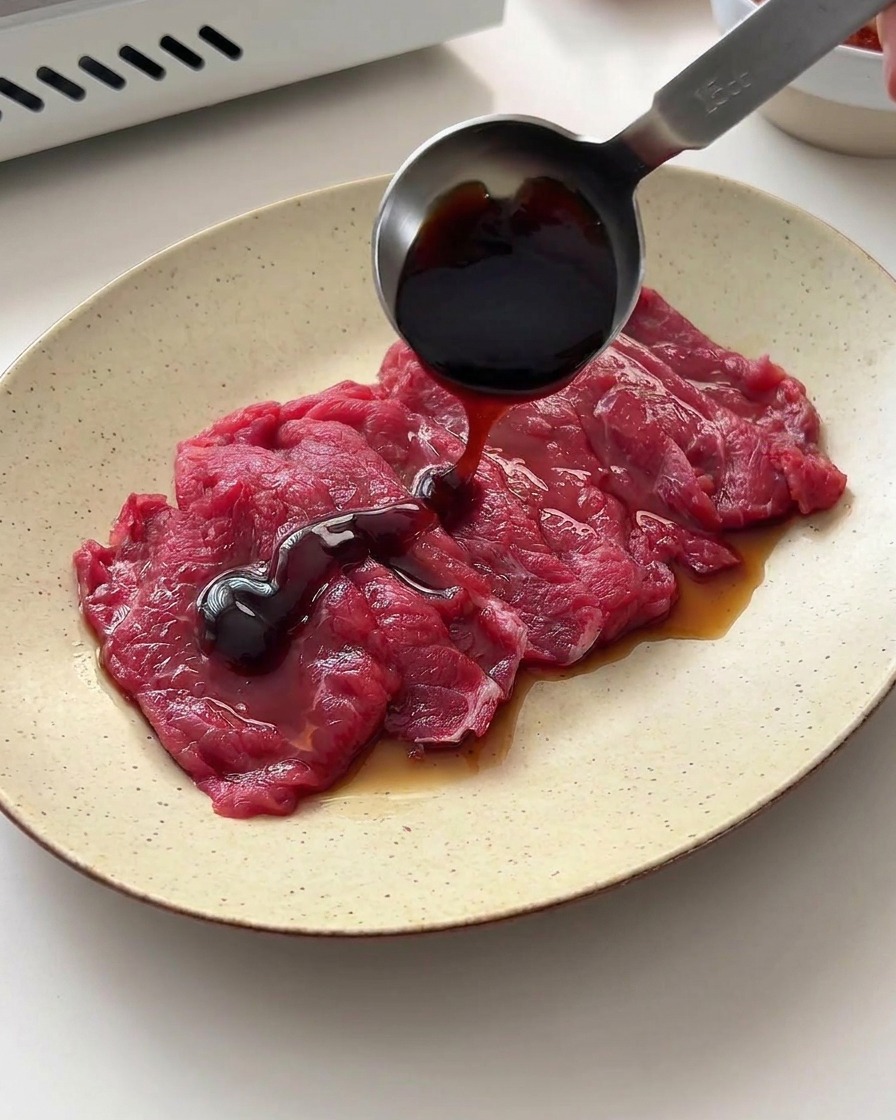 Dark soy sauce and oyster sauce being poured from a metal measuring spoon over raw beef slices on a plate.