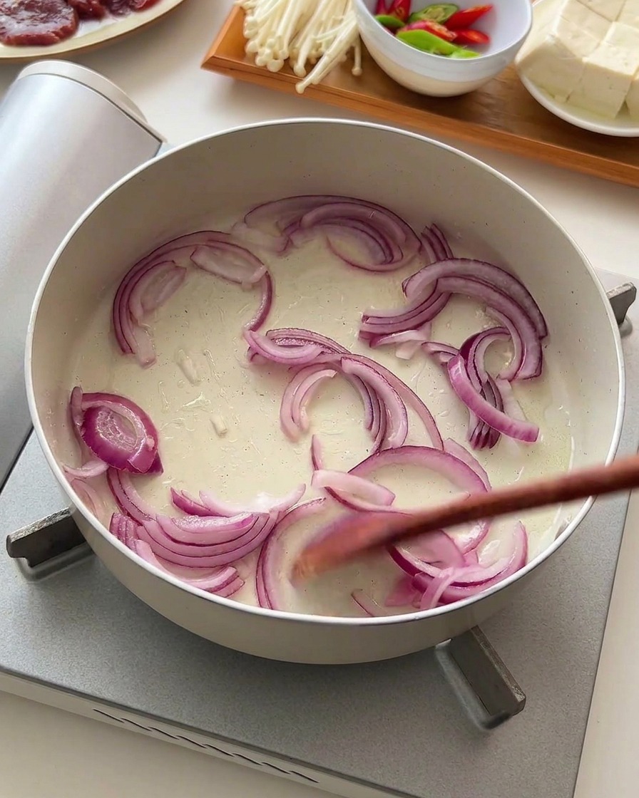 Sliced red onions being stirred with a wooden utensil in a shallow white pan.