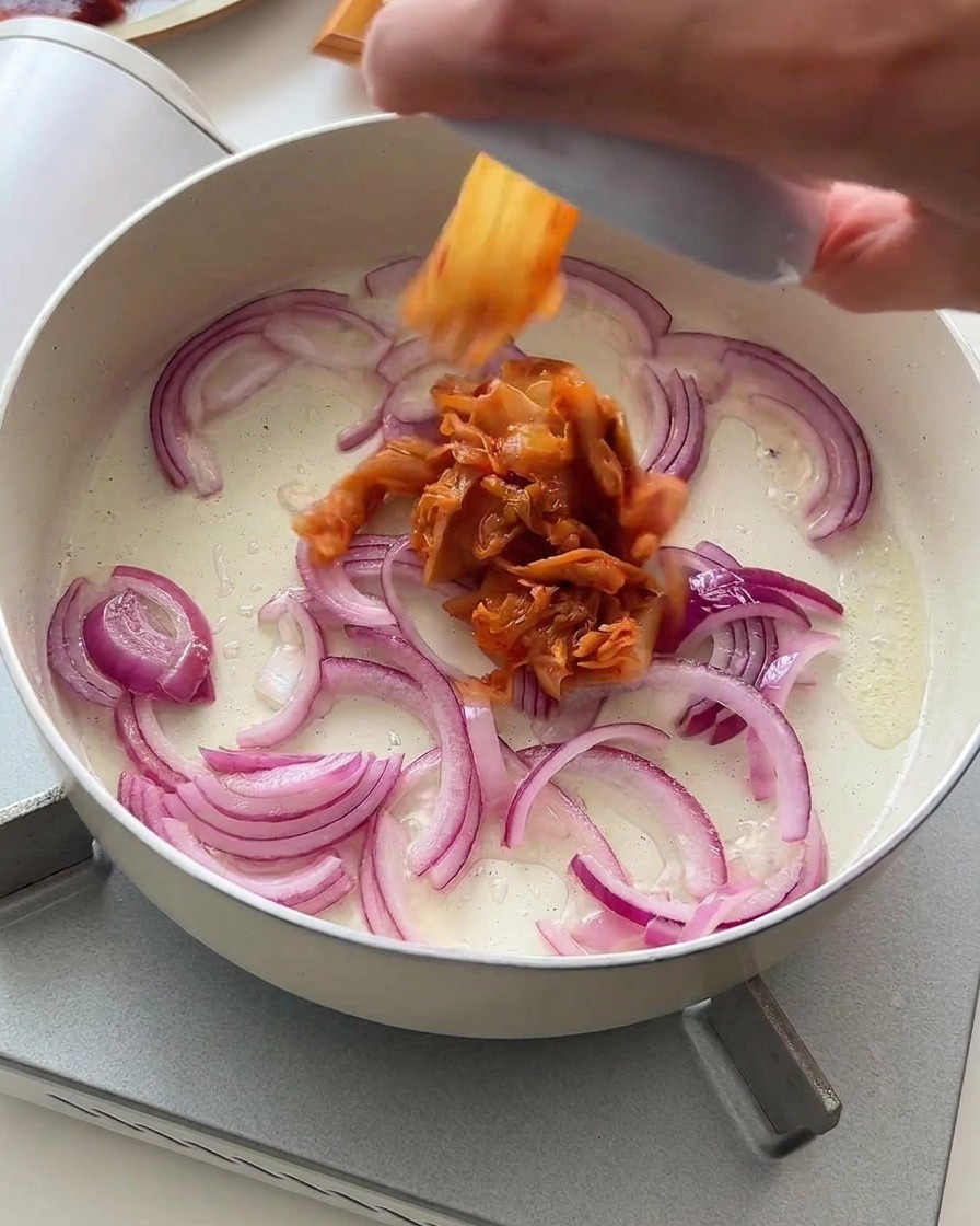 Chopped kimchi being dropped into a pan of sliced red onions gently frying in oil.