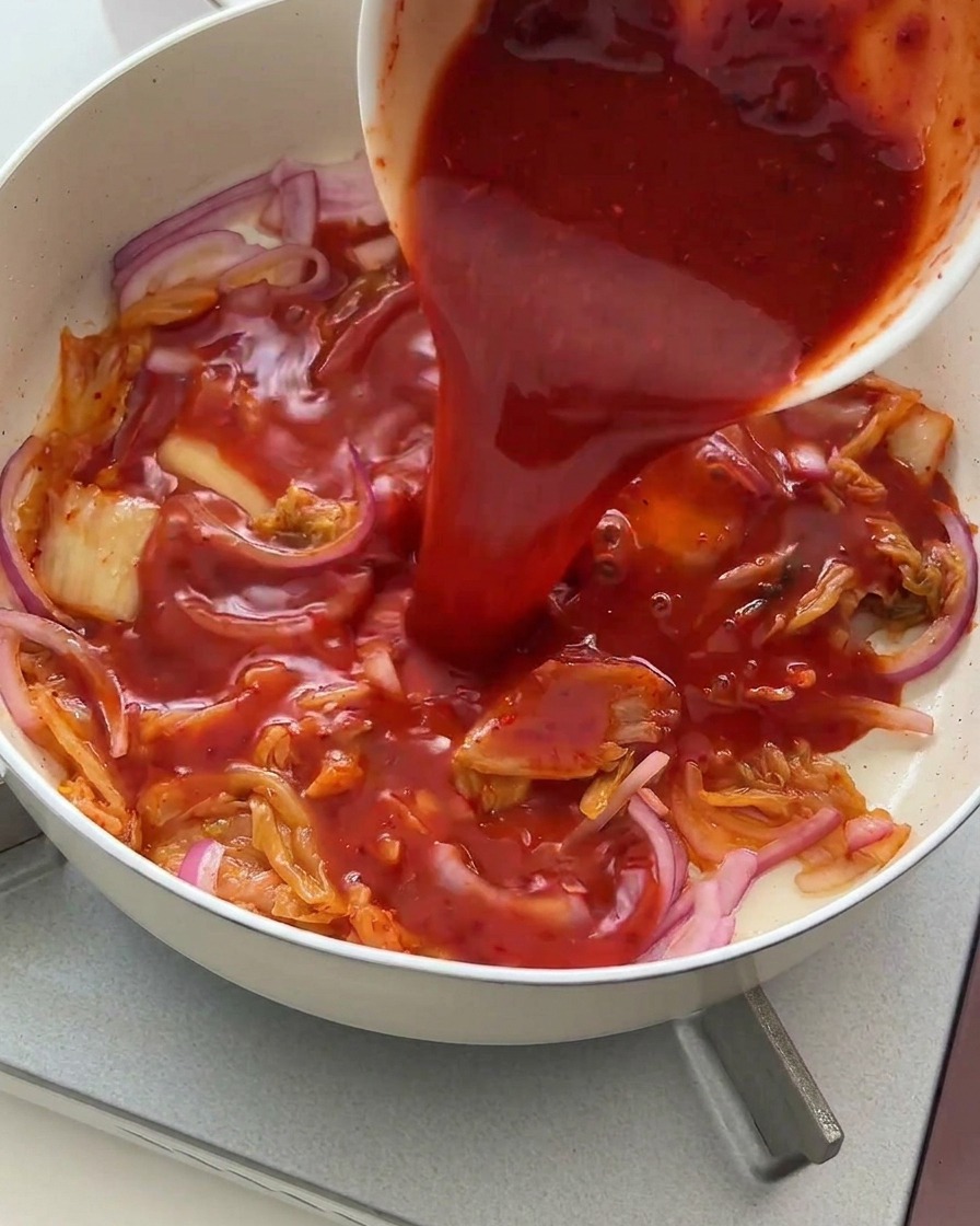 A rich, vibrant red sauce being poured from a white bowl over stir-fried kimchi and red onions.