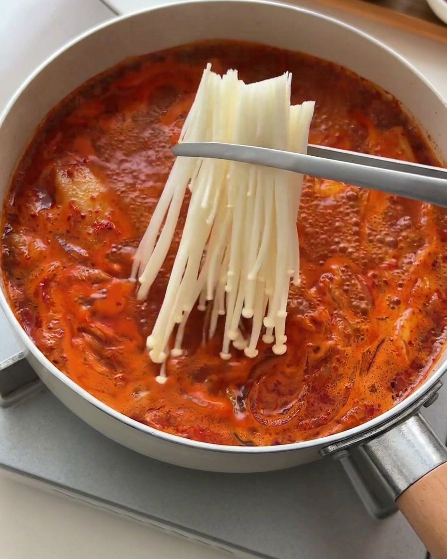 Metal tongs placing a bundle of white, fresh enoki mushrooms into a bubbling, spicy red soup.