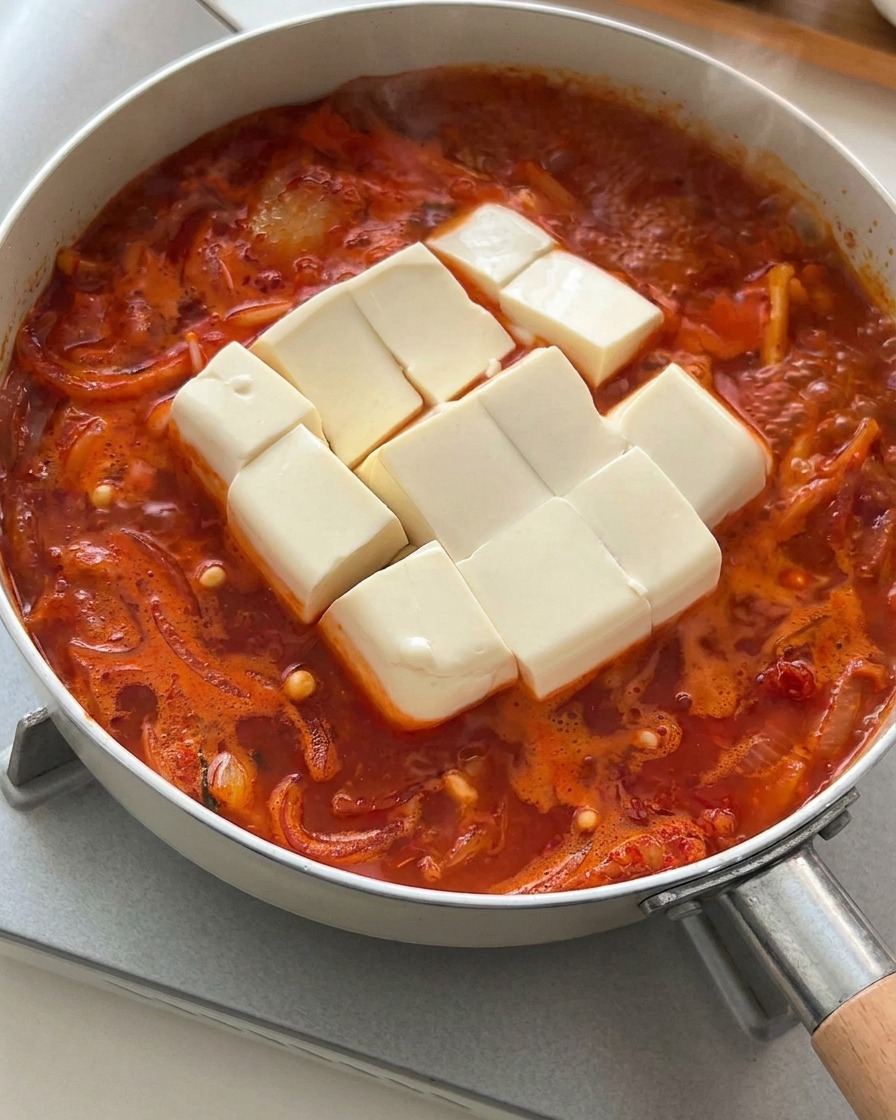 A thick block of soft white tofu, scored into squares, placed gently into a boiling red kimchi stew.