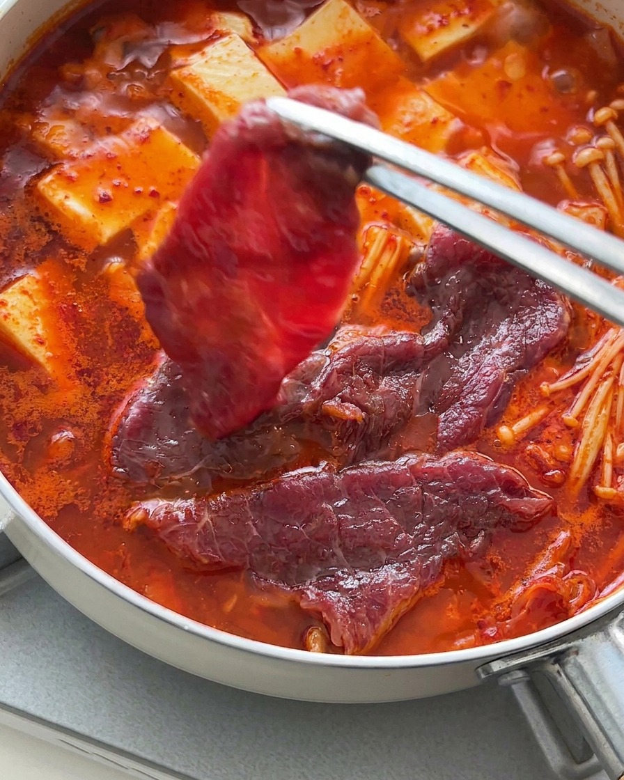Tongs carefully laying thin slices of raw red beef over the hot, bubbling tofu and kimchi broth.