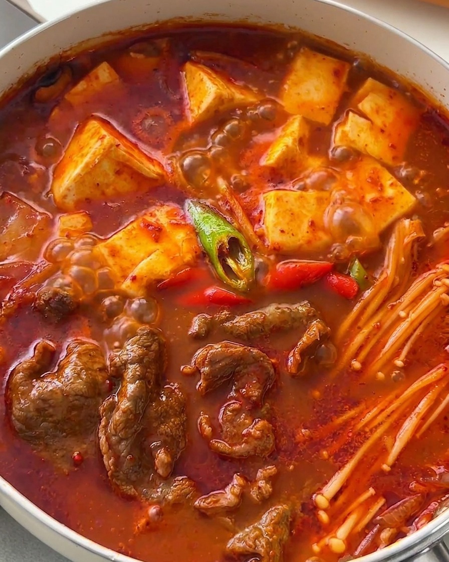 A hearty, bubbling pan of Korean beef and tofu stew brightly garnished with diagonally sliced green and red chili peppers.