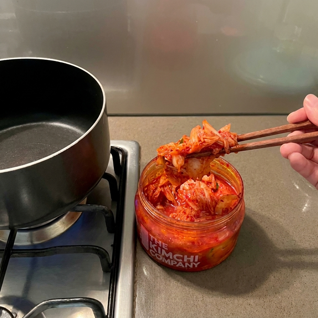 A hand using wooden chopsticks to pull vibrant red kimchi from a jar over an empty black cooking pot.