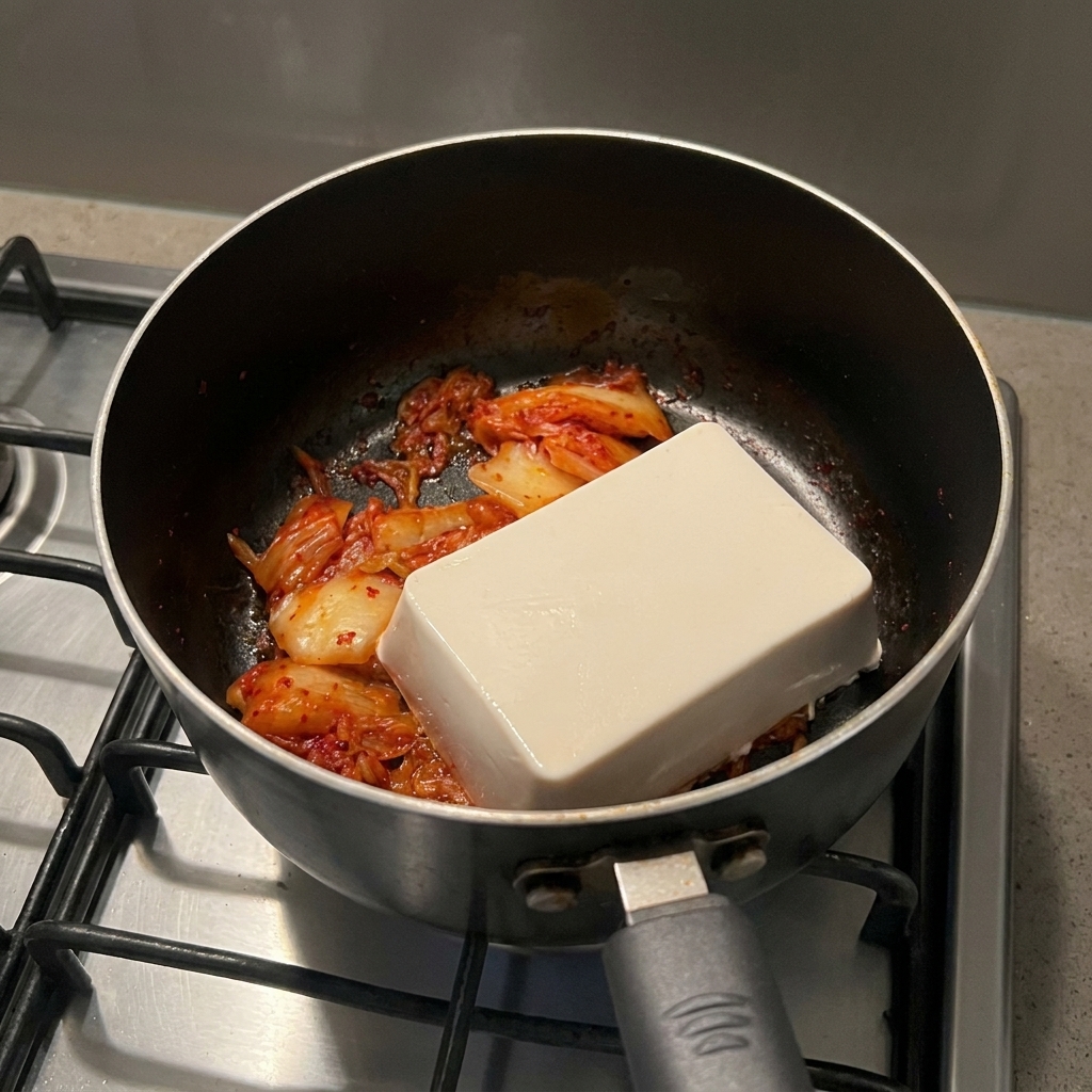 A solid rectangular block of white soft tofu sitting on top of cooked red kimchi in a black cooking pot.