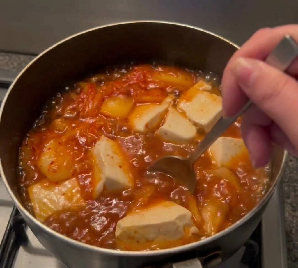 A metal spoon stirring deep red chili paste into a bubbling pot of soup containing soft tofu and kimchi