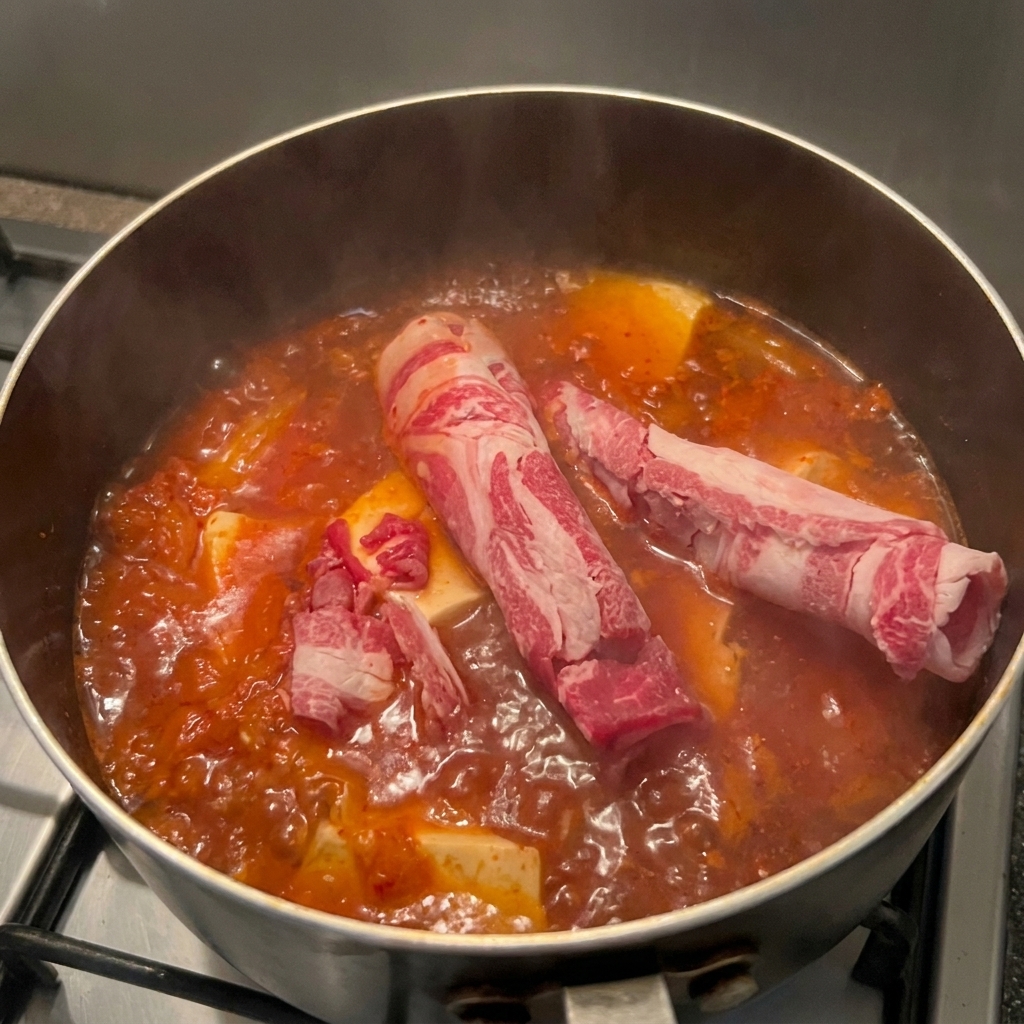 Thinly sliced, marbled raw beef rolls being placed into a pot of boiling red kimchi soup with tofu blocks.