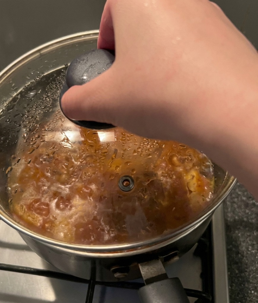 A hand holding the black knob of a glass lid and placing it onto a steaming pot of red stew.