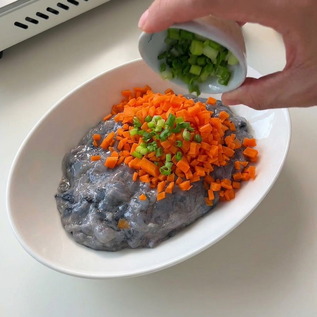 Finely diced orange carrots being poured into a white bowl containing a translucent, grayish raw shrimp paste.