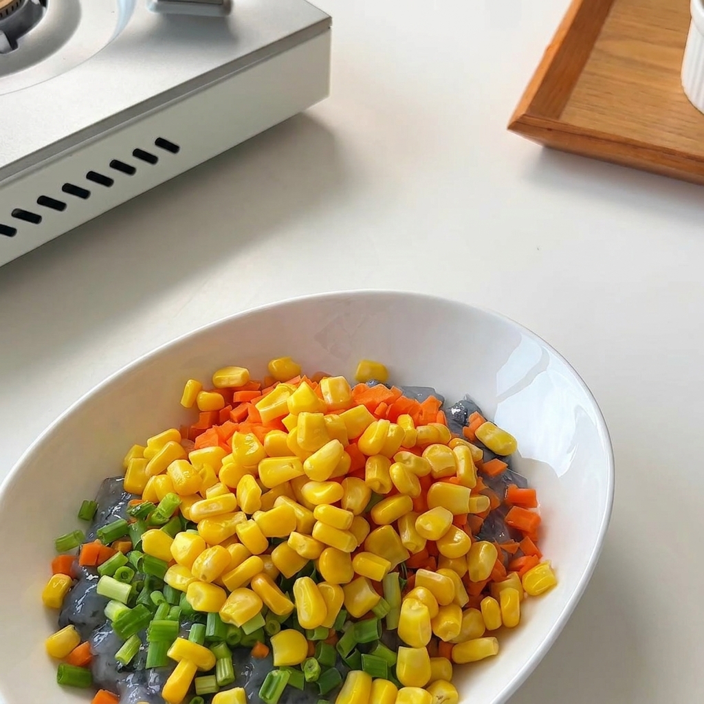 A small bowl of golden sweet corn kernels being emptied into a white mixing bowl with shrimp paste, carrots, and scallions.