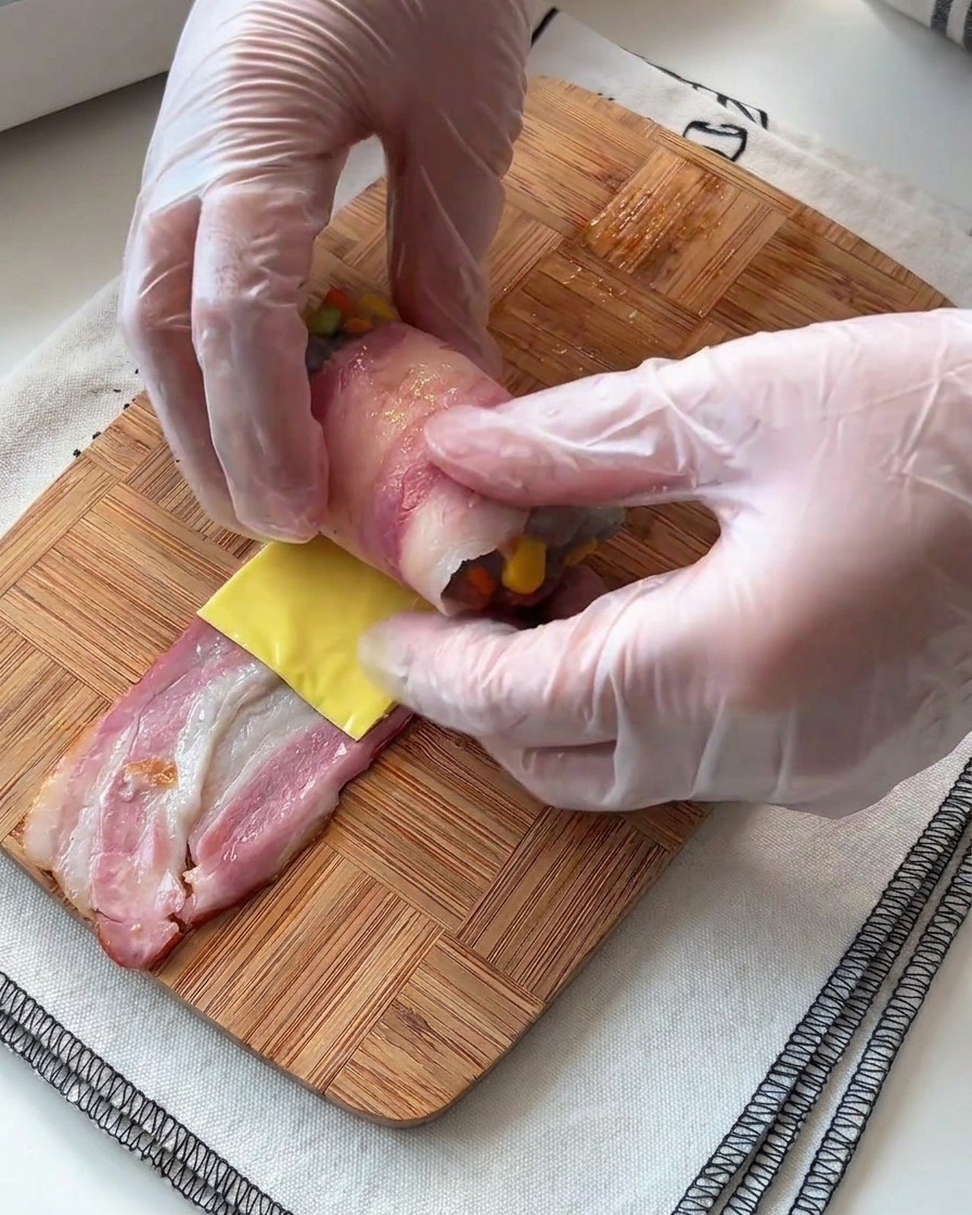 Hands carefully rolling a strip of bacon around a shrimp and cheese filling on a wooden surface.