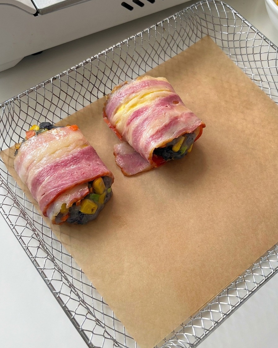 Two raw bacon-wrapped shrimp rolls sitting on brown parchment paper inside a wire air fryer basket.
