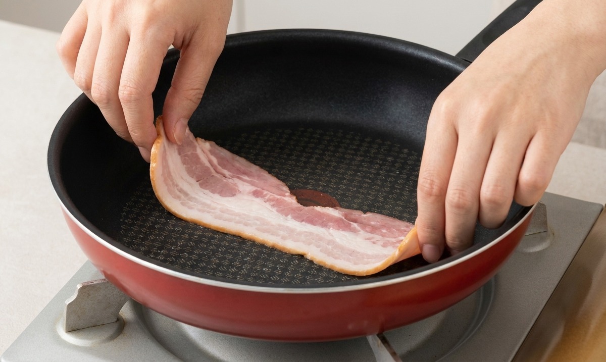 Hands placing raw strips of bacon into a preheated, lightly oiled frying pan.
