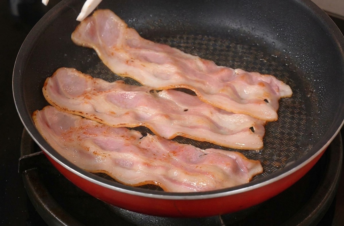 Tongs flipping partially cooked, curling bacon strips in a red frying pan on a stovetop.