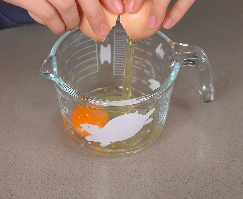 Hands cracking a brown egg into a glass cup that already contains one raw egg.