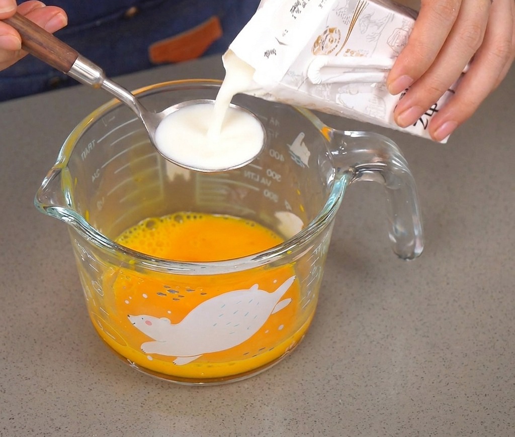 Pouring milk from a carton into a silver spoon over a cup of whisked eggs.