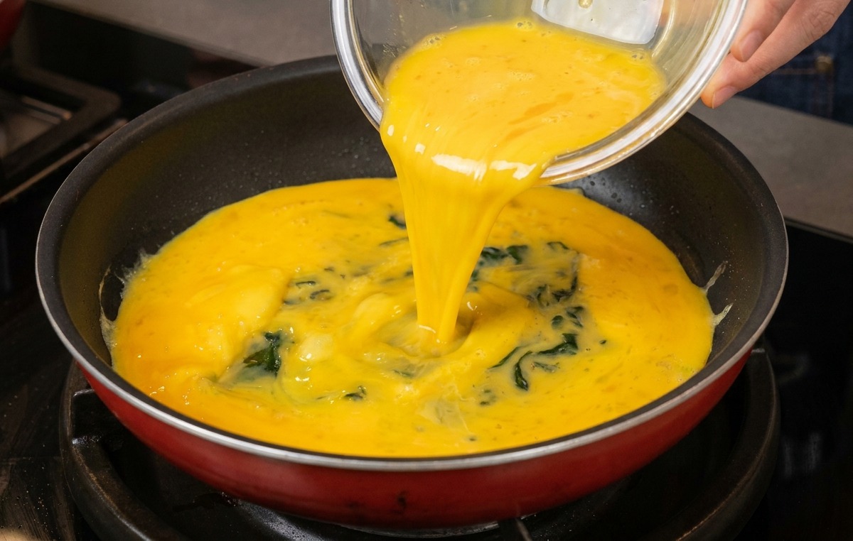 Yellow beaten egg mixture being poured from a glass bowl into a clean frying pan