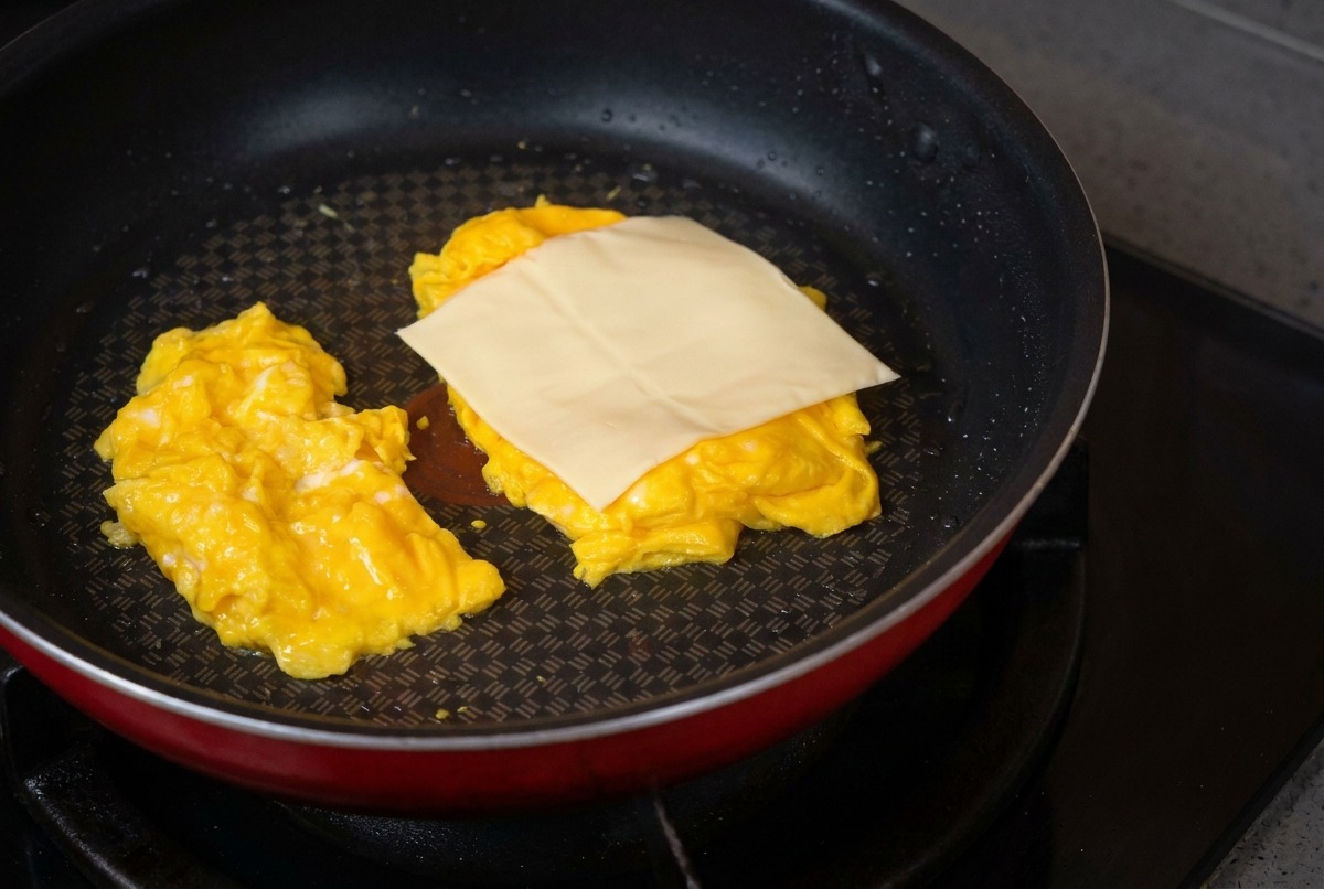 A non-stick frying pan containing two mounds of soft scrambled eggs, with a pale square slice of cheese being placed on top of one mound.