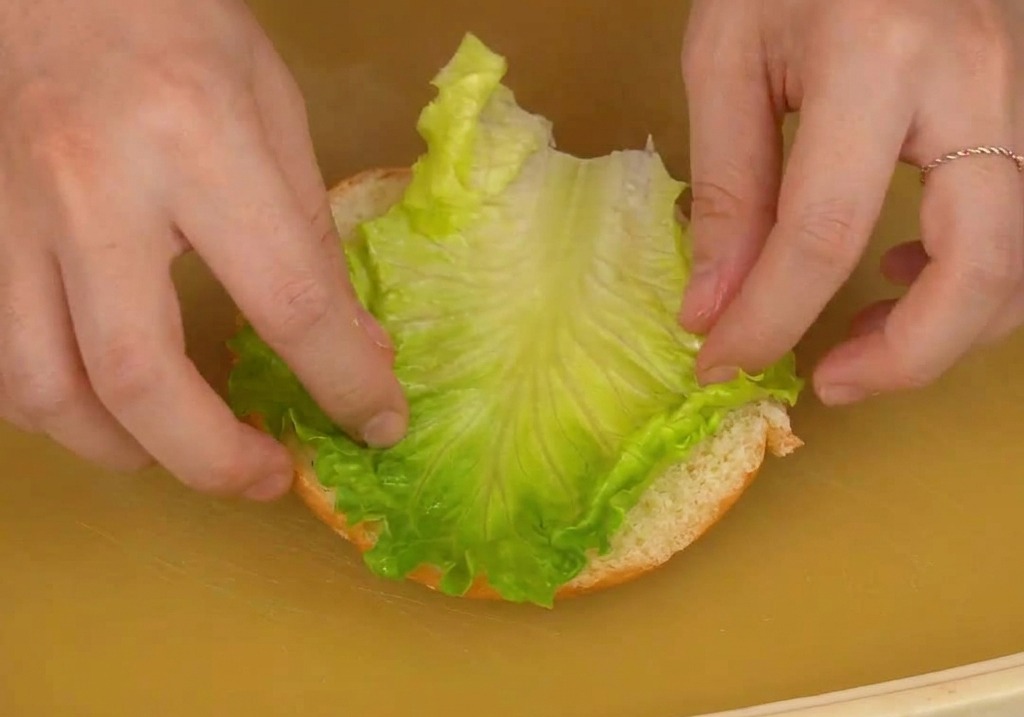 Two hands carefully placing a large, fresh leaf of green lettuce onto the bottom half of a burger bun on a cutting board.