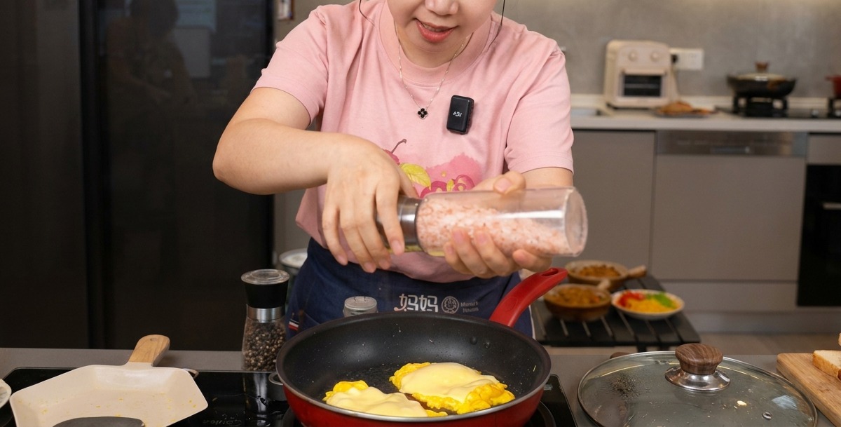 A person grinding sea salt and black pepper from a transparent mill onto fluffy scrambled eggs topped with a layer of melted cheese in a red frying pan.