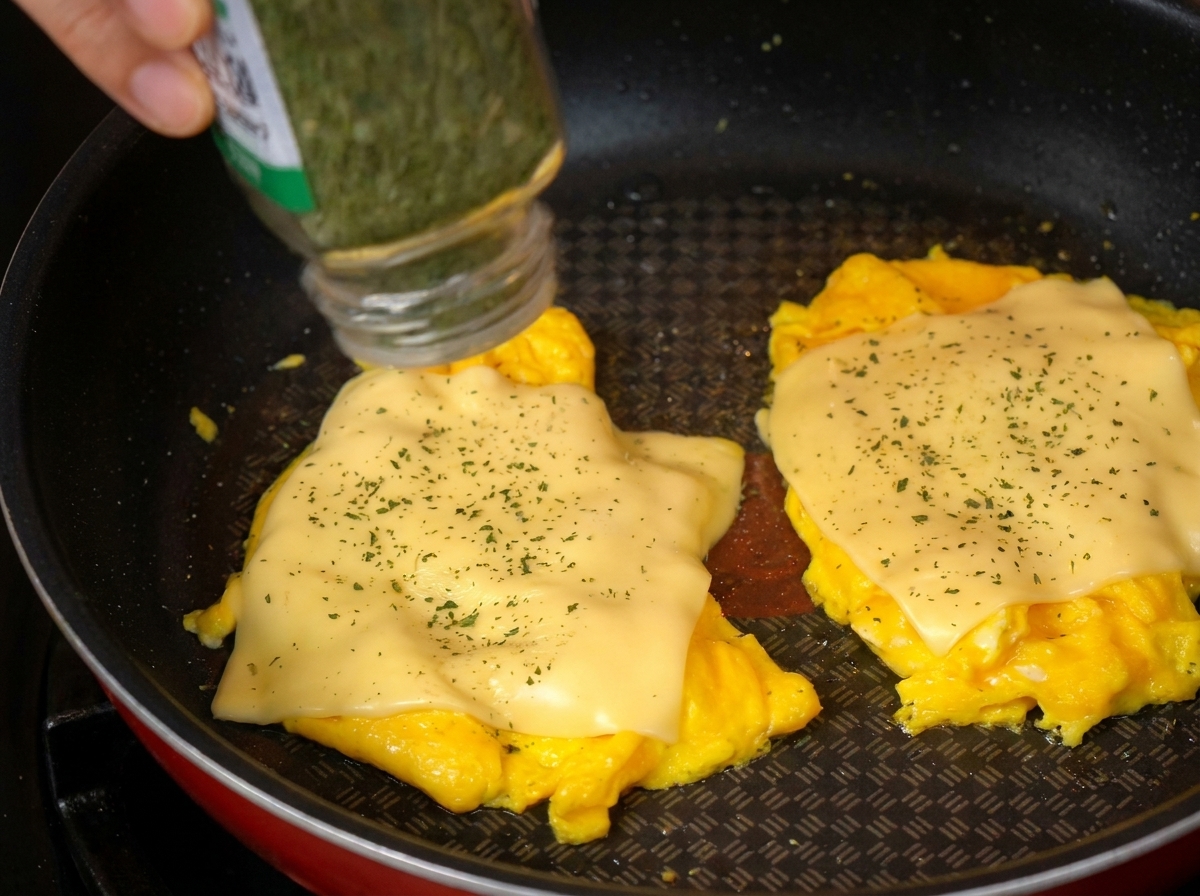 A close-up of dried parsley being sprinkled from a spice container onto scrambled eggs topped with melted cheese in a pan.