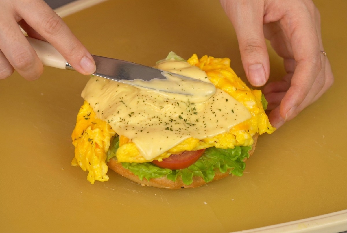 A close-up shot of a person using a spatula to place a portion of cooked scrambled eggs with melted cheese onto a burger bun layered with lettuce and tomato.