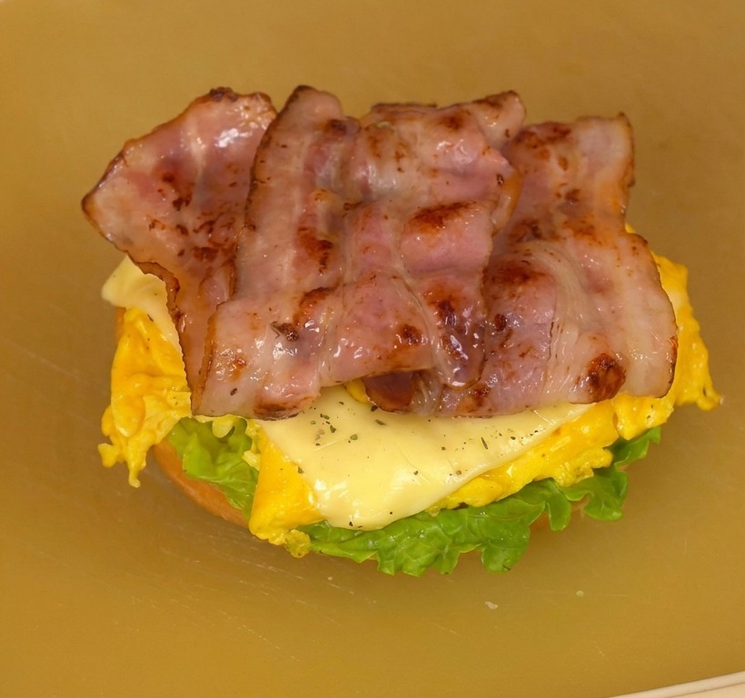 An open burger assembly on a cutting board showing layers of fresh lettuce, tomato slices, cheesy scrambled eggs, and crispy bacon.