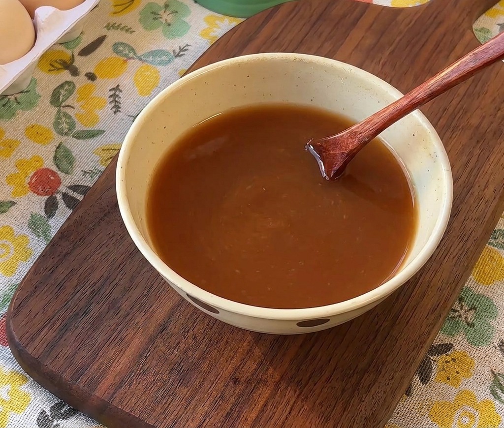 A wooden spoon stirring a brown liquid sauce mixture in a small ceramic bowl.