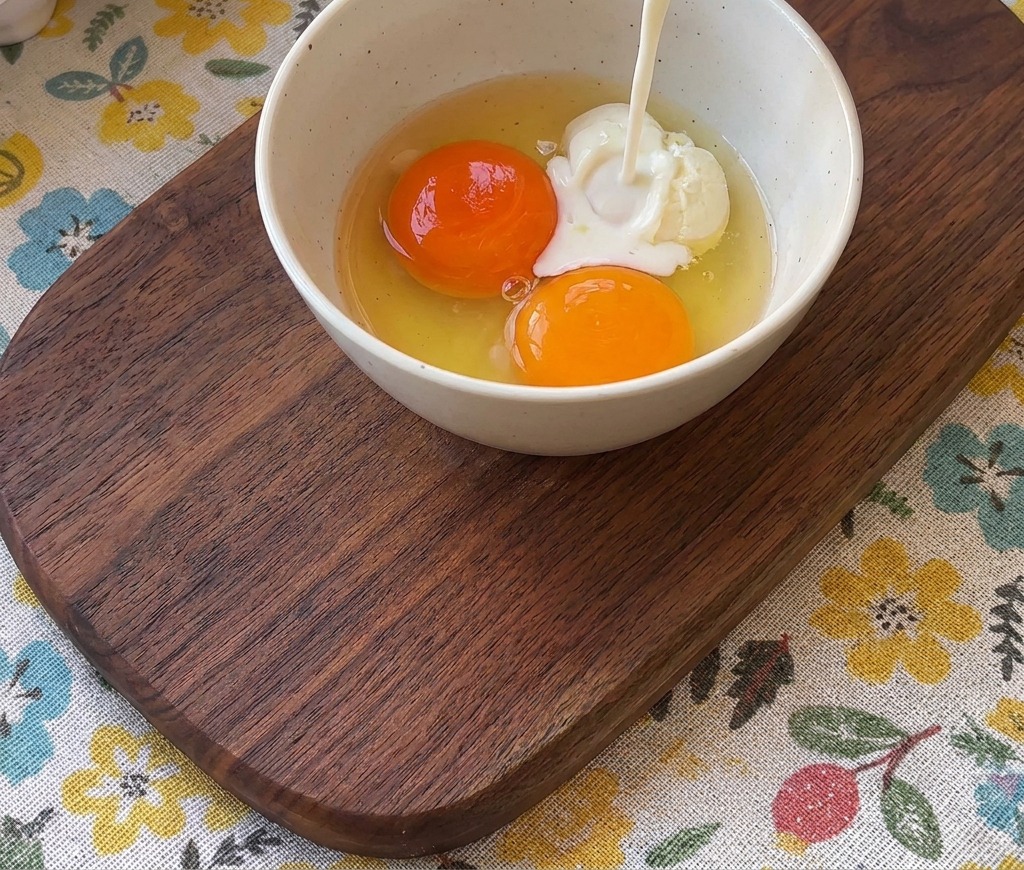 Milk being poured from a glass into a bowl containing two bright orange egg yolks.