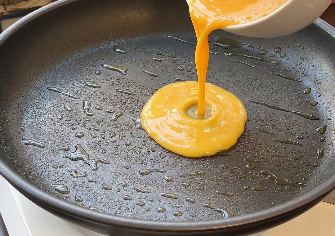 Yellow beaten egg mixture being poured from a white bowl into a black frying pan with glistening oil.