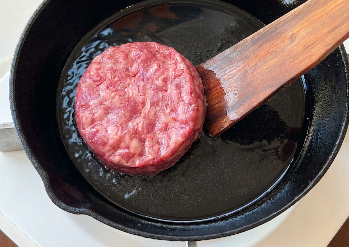 A raw ground beef patty being placed into a preheated cast-iron skillet with a wooden spatula.