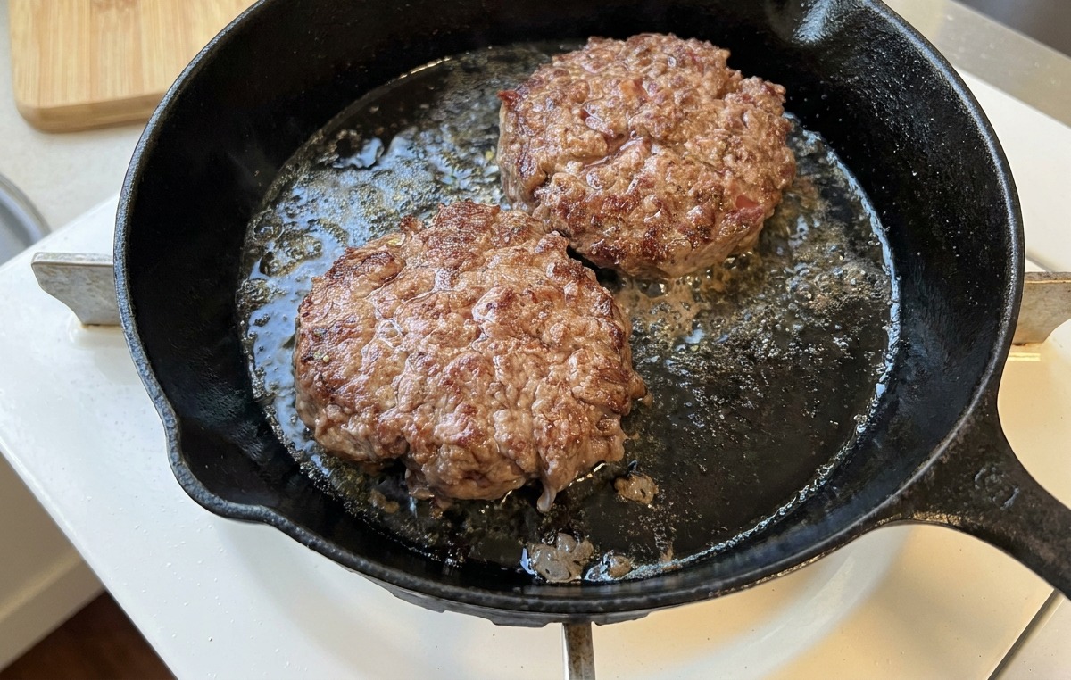 Two beef patties sizzling in a cast-iron skillet, showing a deeply browned and caramelized crust.