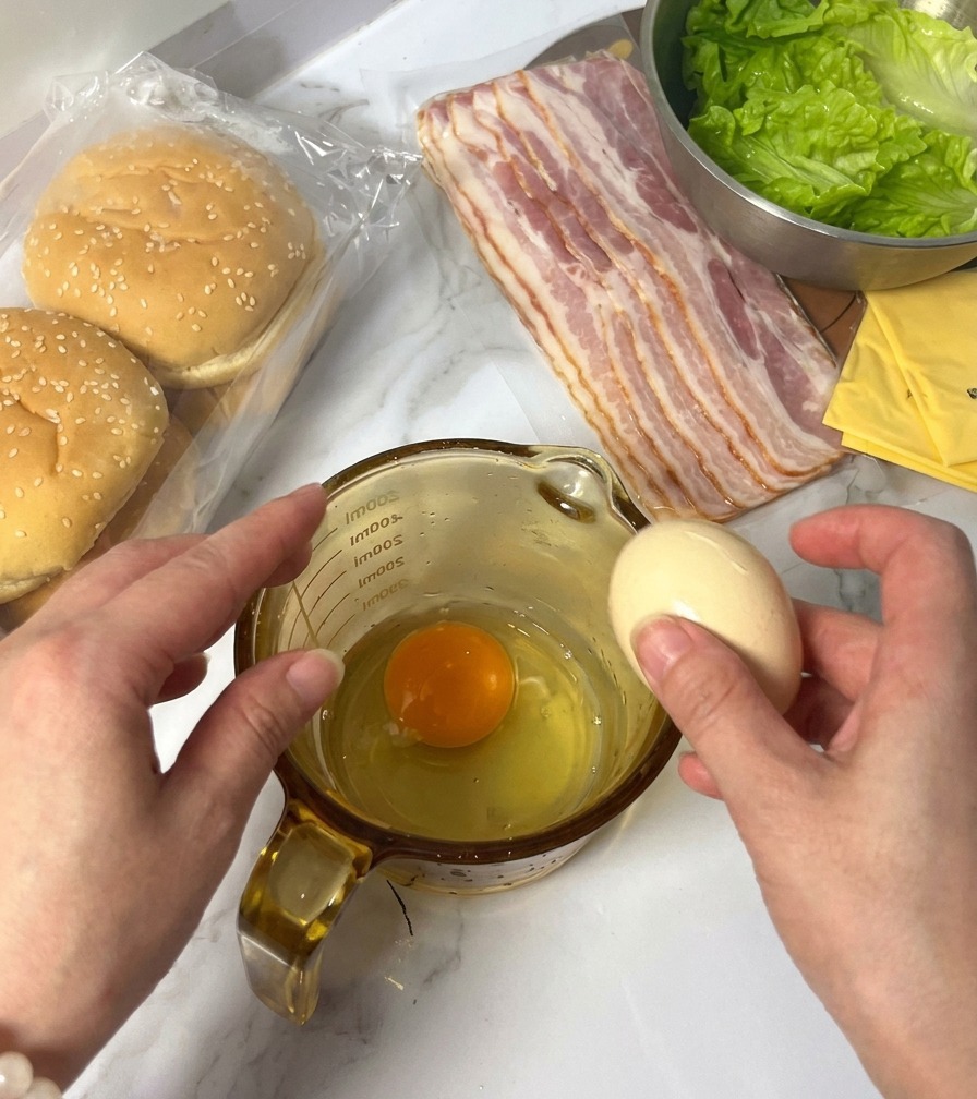 A person cracking a raw egg into a glass measuring cup that already contains one egg yolk and white, with burger ingredients visible in the background.