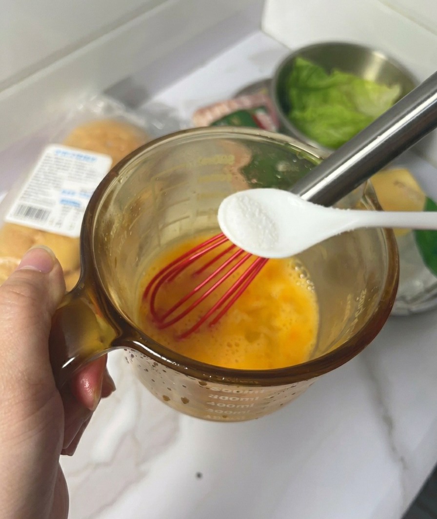 A hand holding a small white spoon filled with salt over a glass cup containing whisked yellow eggs and a red whisk.