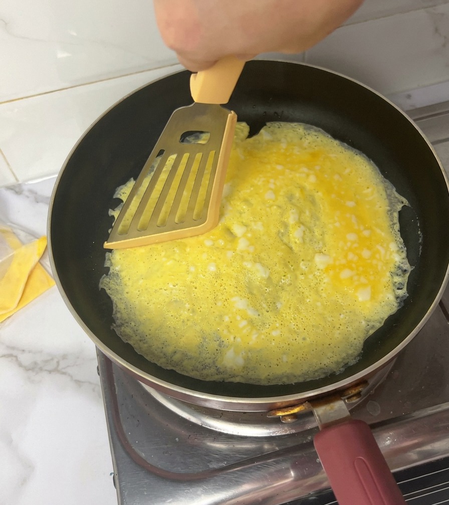 A hand using a yellow spatula to slice a thin yellow omelet into four sections inside a black frying pan.