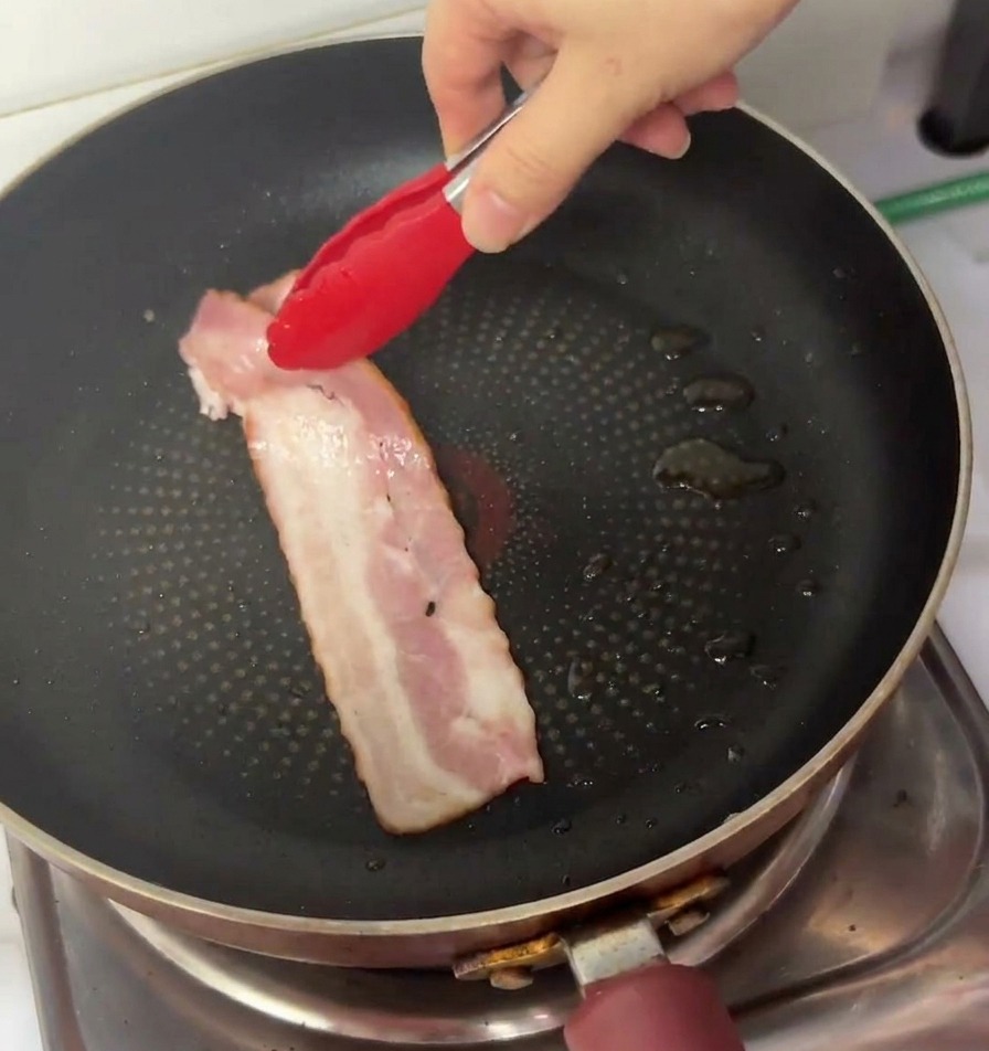 A hand using red tipped tongs to place a long strip of raw pink bacon into a black non stick frying pan.