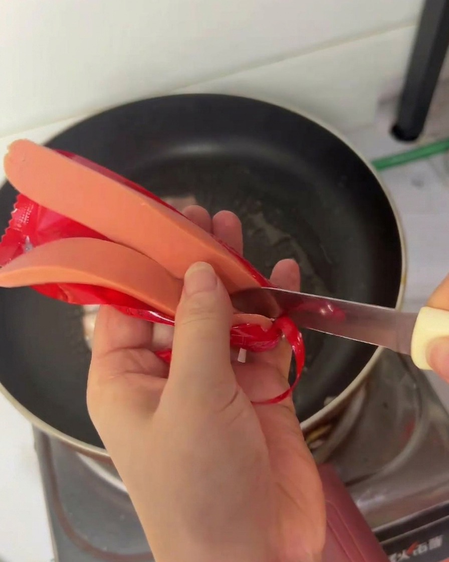 A person using a small knife to slice a red-cased hot dog sausage in half lengthwise over a frying pan.